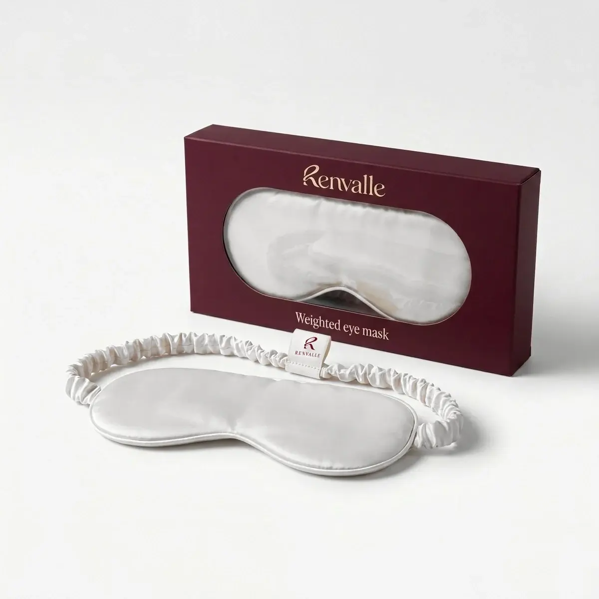 The Weighted Eye Mask in Ivory with its gathered strap, in front of its burgundy box with a window, the design target