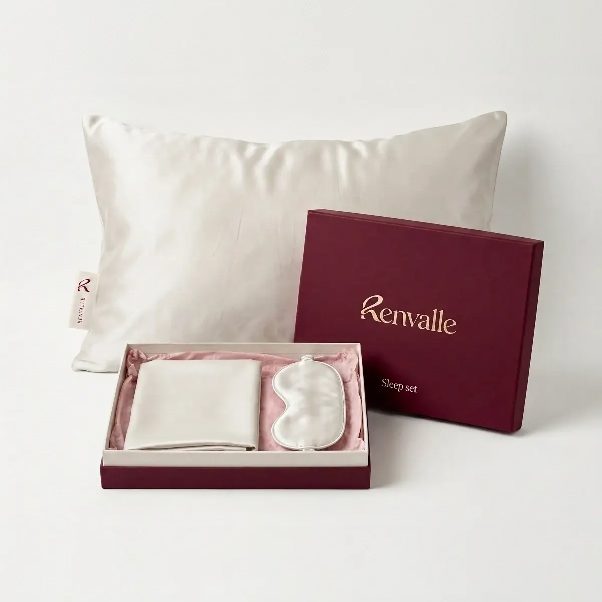 The Sleep Set in Ivory: the pillowcase standing on a pillow, the burgundy set box open in front with a folded pillowcase and the eye mask inside