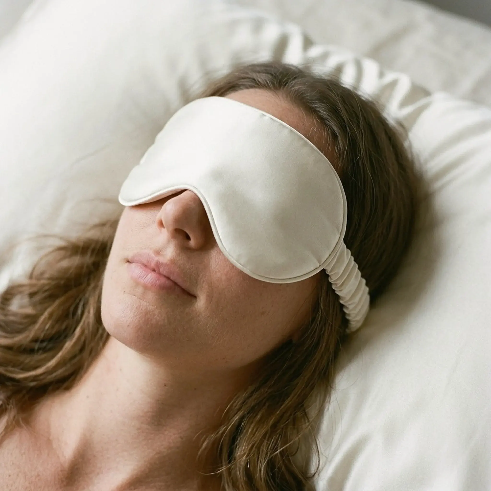 A woman lying back on a silk pillowcase wearing the Weighted Eye Mask in Ivory