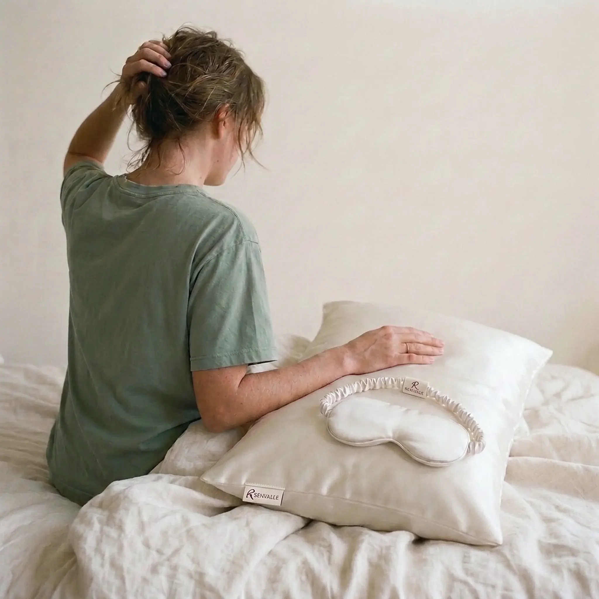 A woman seen from behind on a linen bed, one hand on a pillow in the Ivory silk pillowcase with the Ivory eye mask resting on it